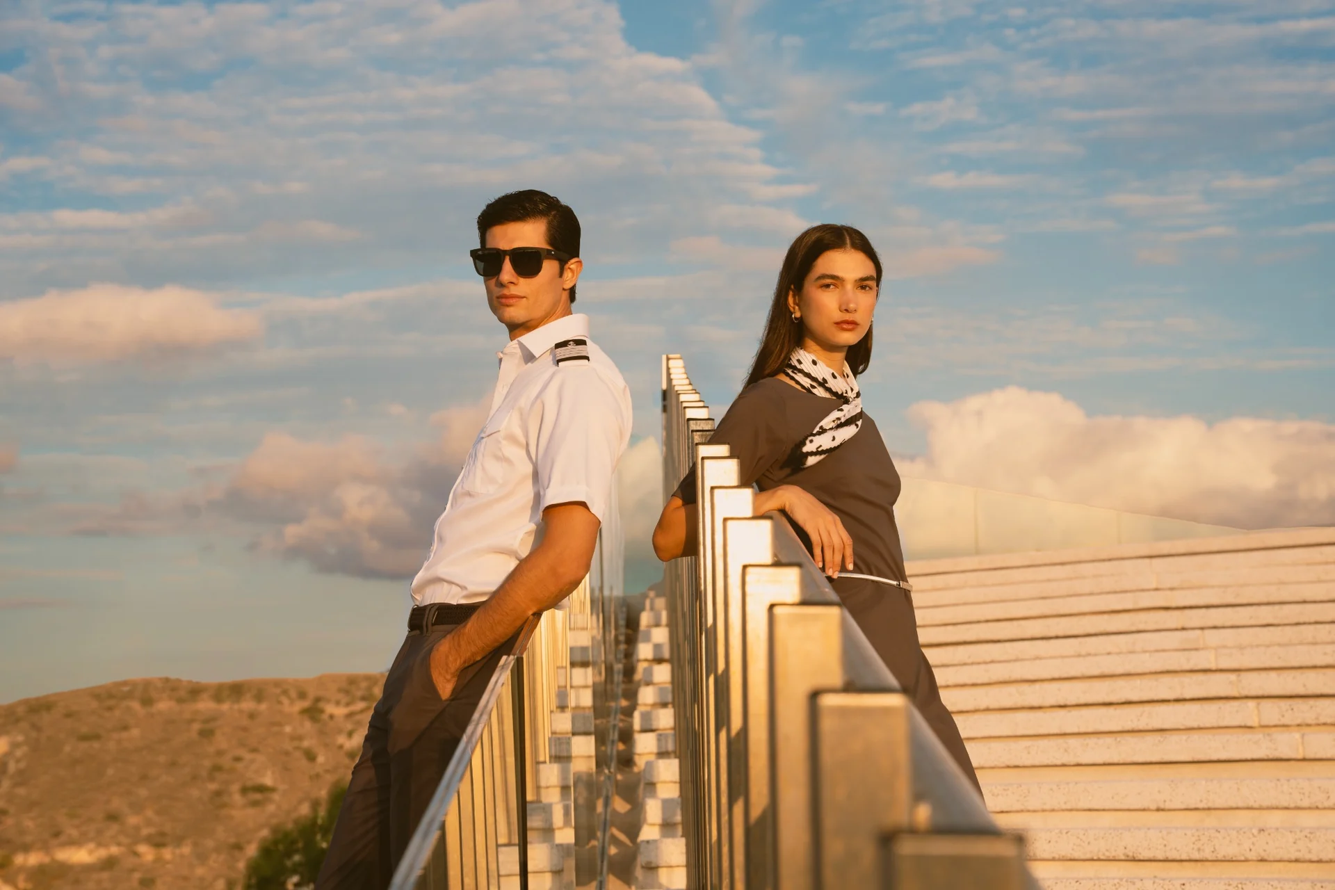 Two models on opposite sides of a stair railing in warm evening light