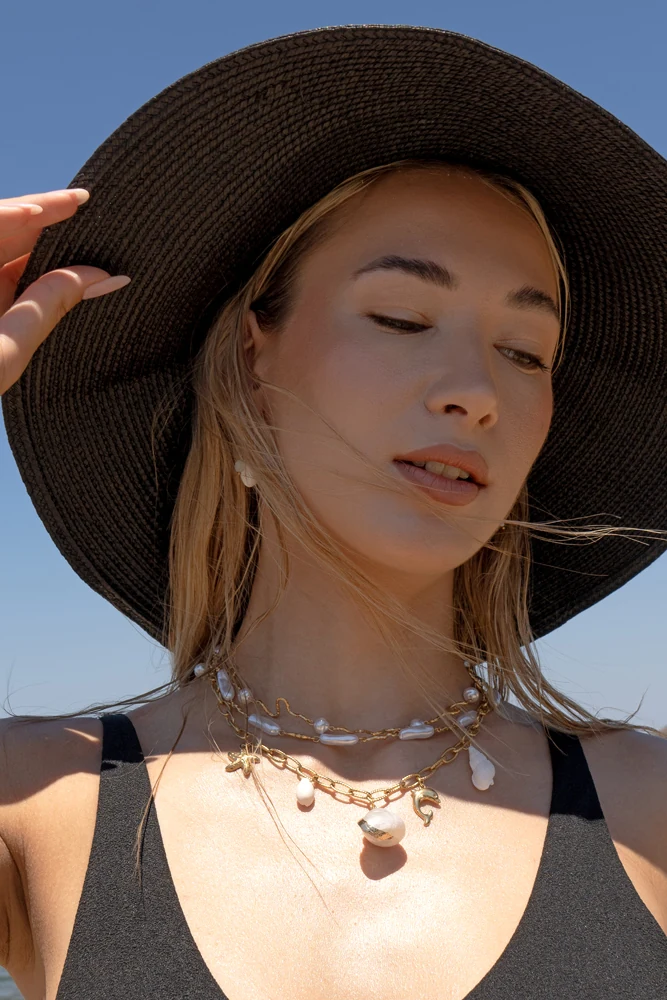 Sunlit portrait beneath a wide black hat, with layered necklaces and shell jewellery