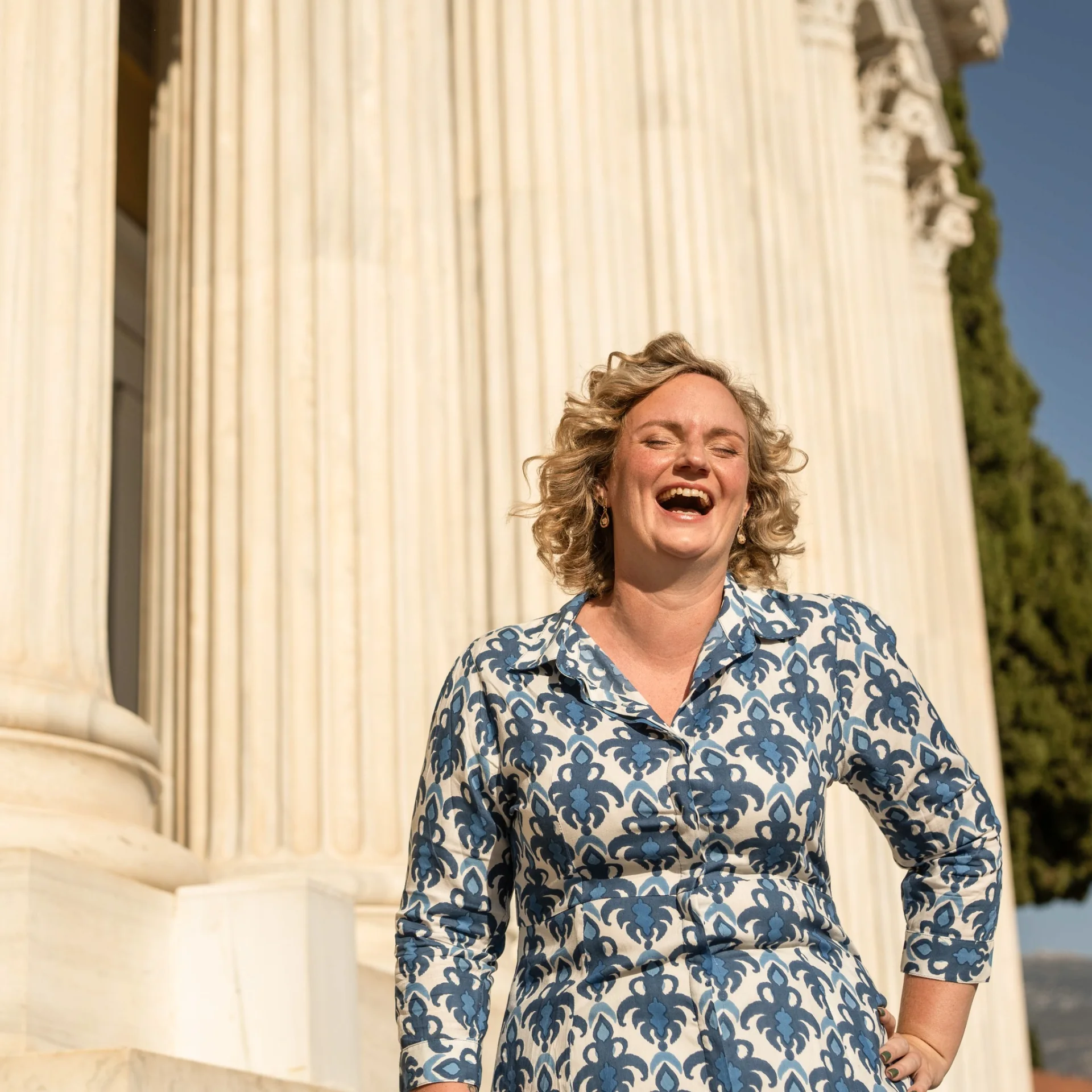 Melina laughing in a blue patterned dress beside classical columns