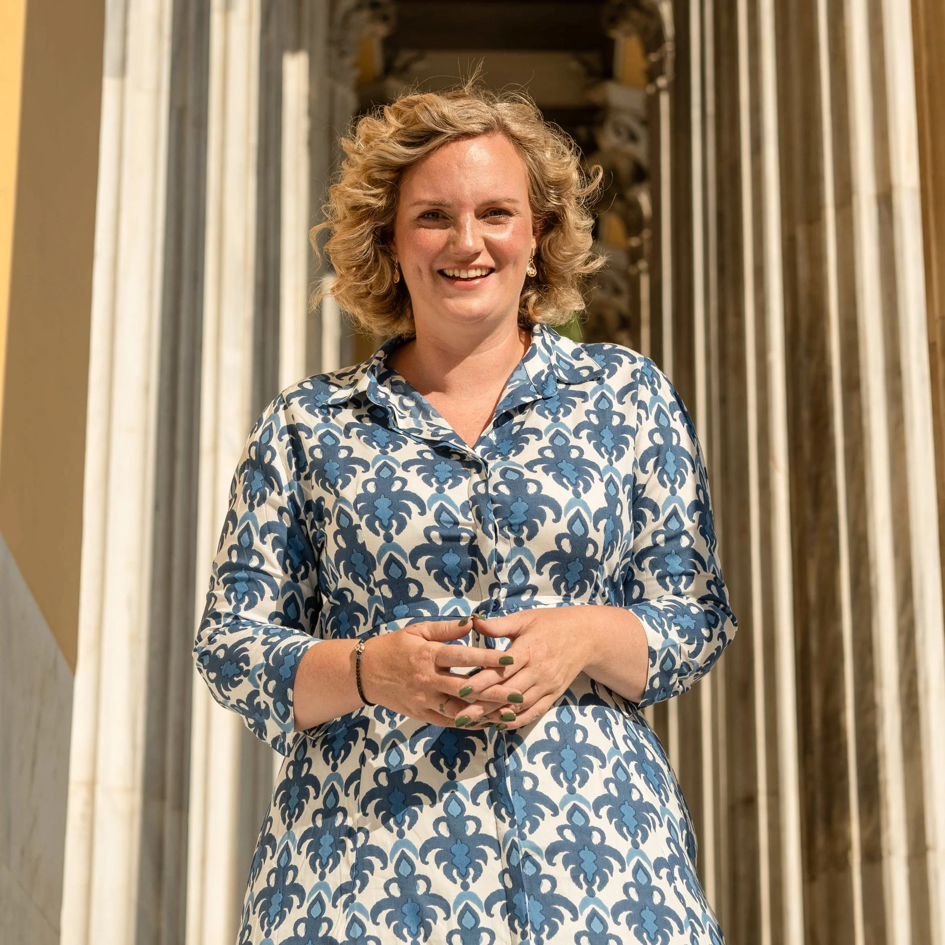 Melina smiling between sunlit columns in a blue patterned dress