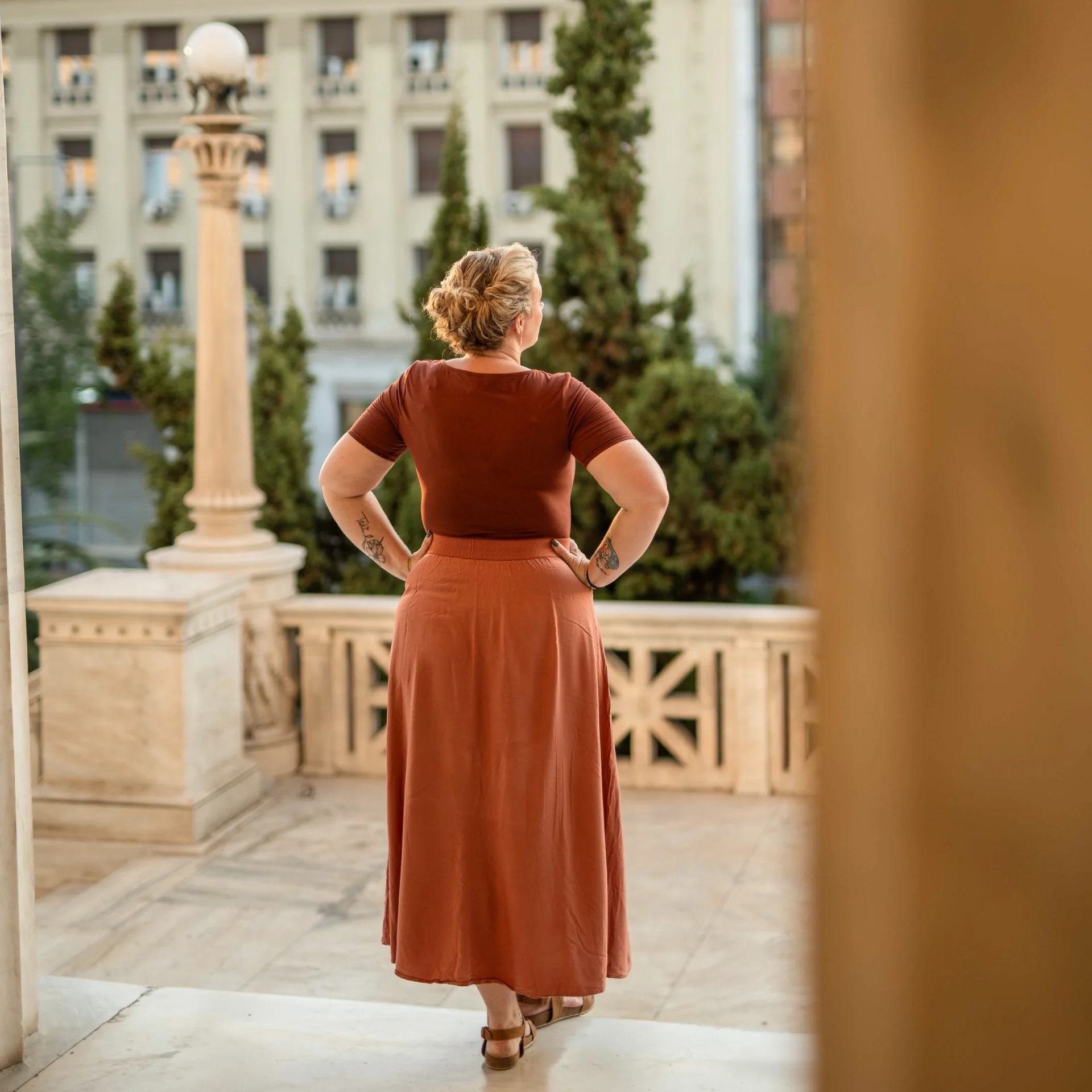 Melina seen from behind, looking towards a green courtyard and Athens buildings