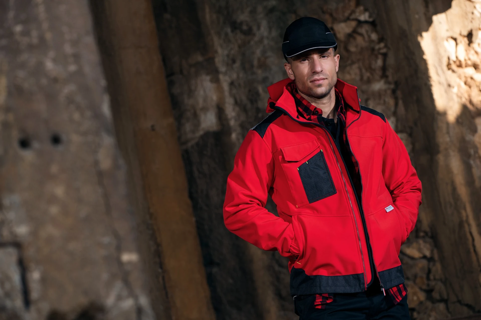 Model in a red and black work jacket and black cap against a textured industrial wall