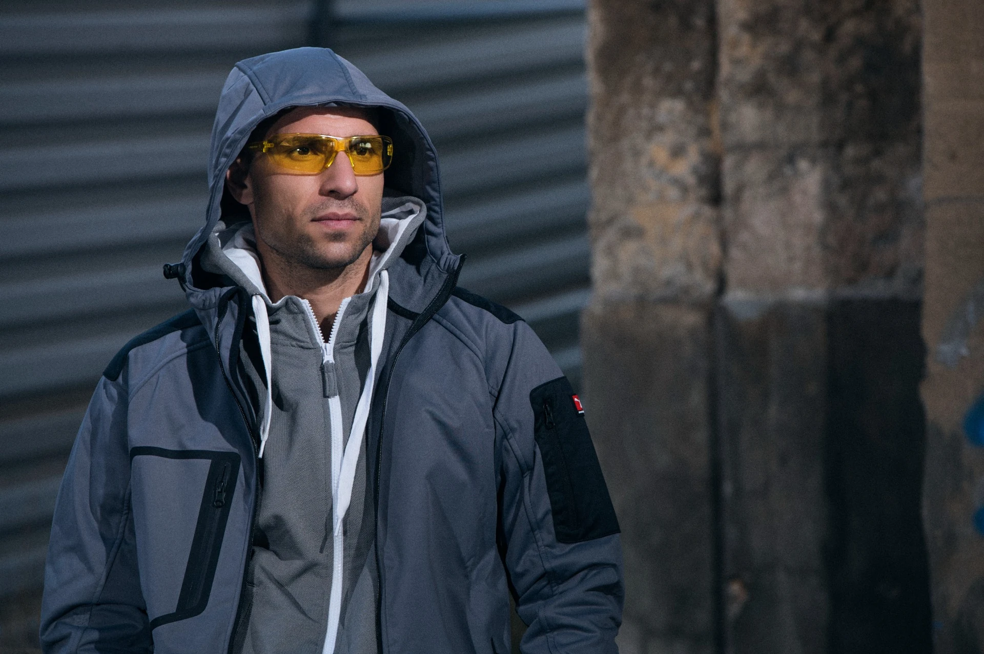 Model in a grey hooded work jacket and yellow protective glasses