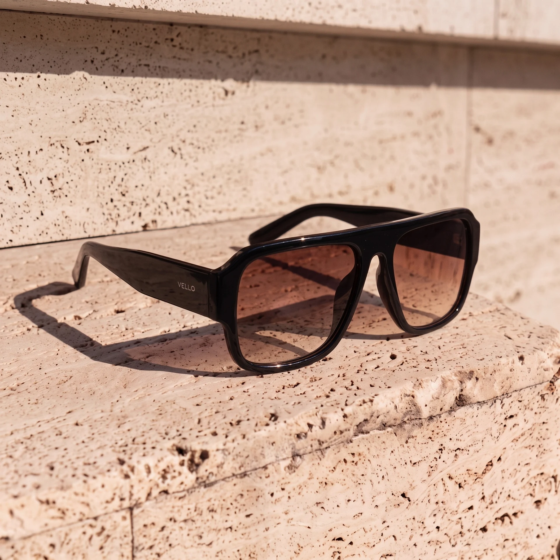 Black Vello sunglasses with gradient lenses and a long shadow on pale stone